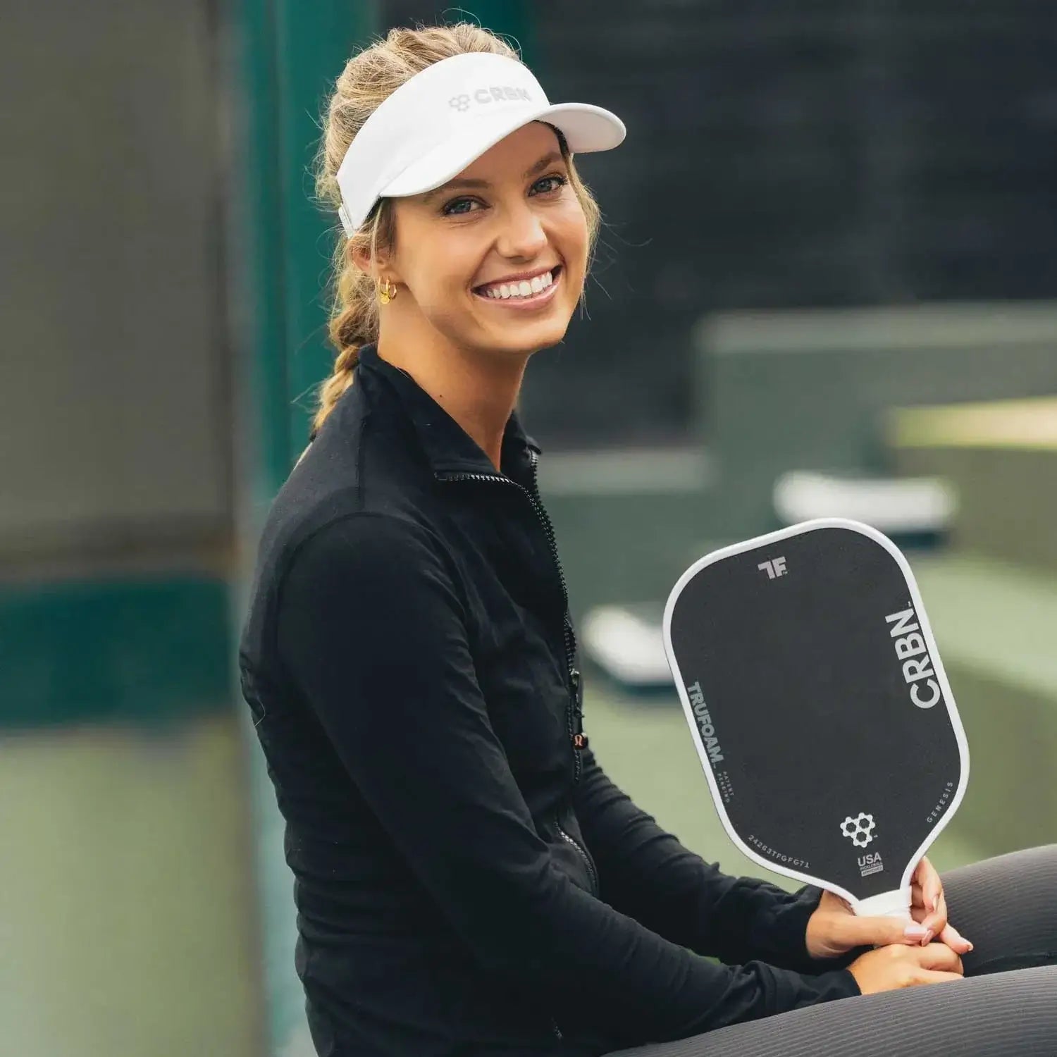 Smiling woman in a visor holding CRBN TruFoam™ paddle, showcasing performance in pickleball.