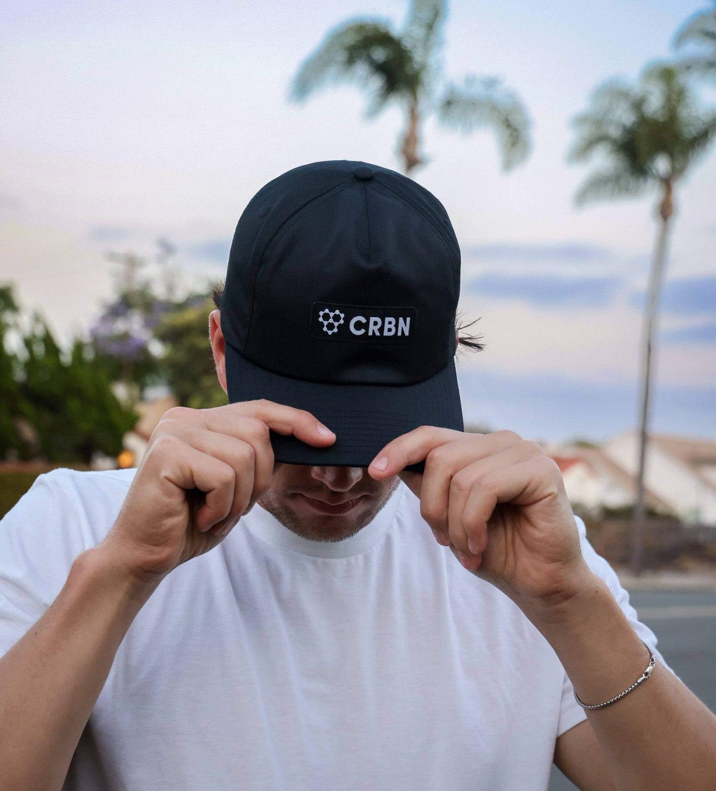 CRBN Essential Snapback Hat worn by a person adjusting the cap against a sunset backdrop with palm trees.