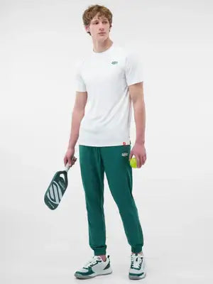 A man in a white short-sleeve performance tee and green joggers, holding a pickleball paddle and ball.