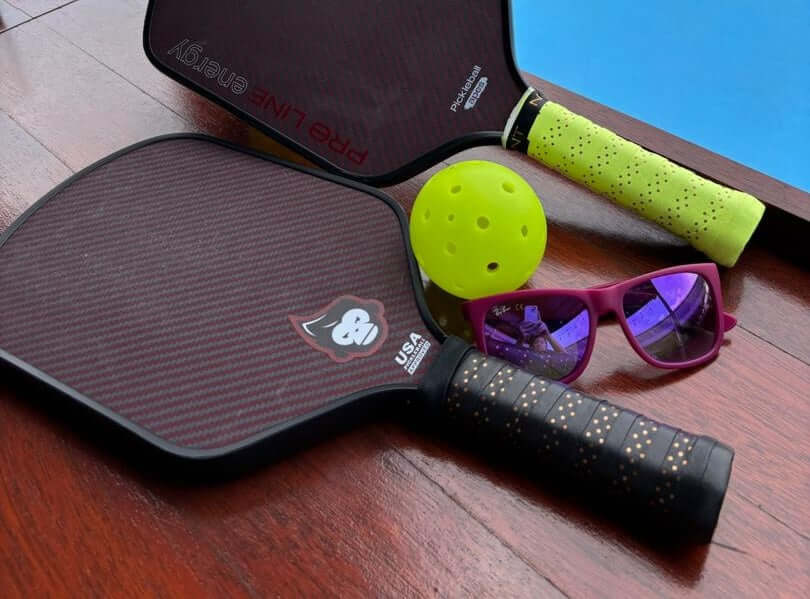 PickleballApes Energy S paddle with yellow pickleball and sunglasses by the court.