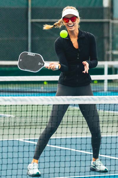 Woman playing pickleball with CRBN glasses on a court, smiling and ready to hit the ball with a paddle.