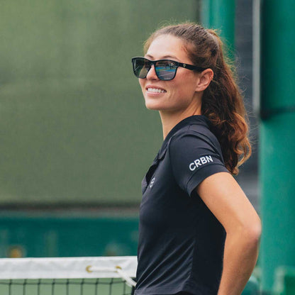 Woman wearing CRBN Drift Pickleball sunglasses, smiling, in a sporty setting, showcasing stylish black frames.