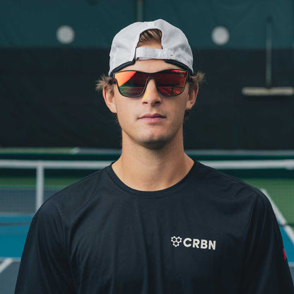 Young man in CRBN Drift Pickleball glasses with reflective lenses, wearing a black shirt on a tennis court.