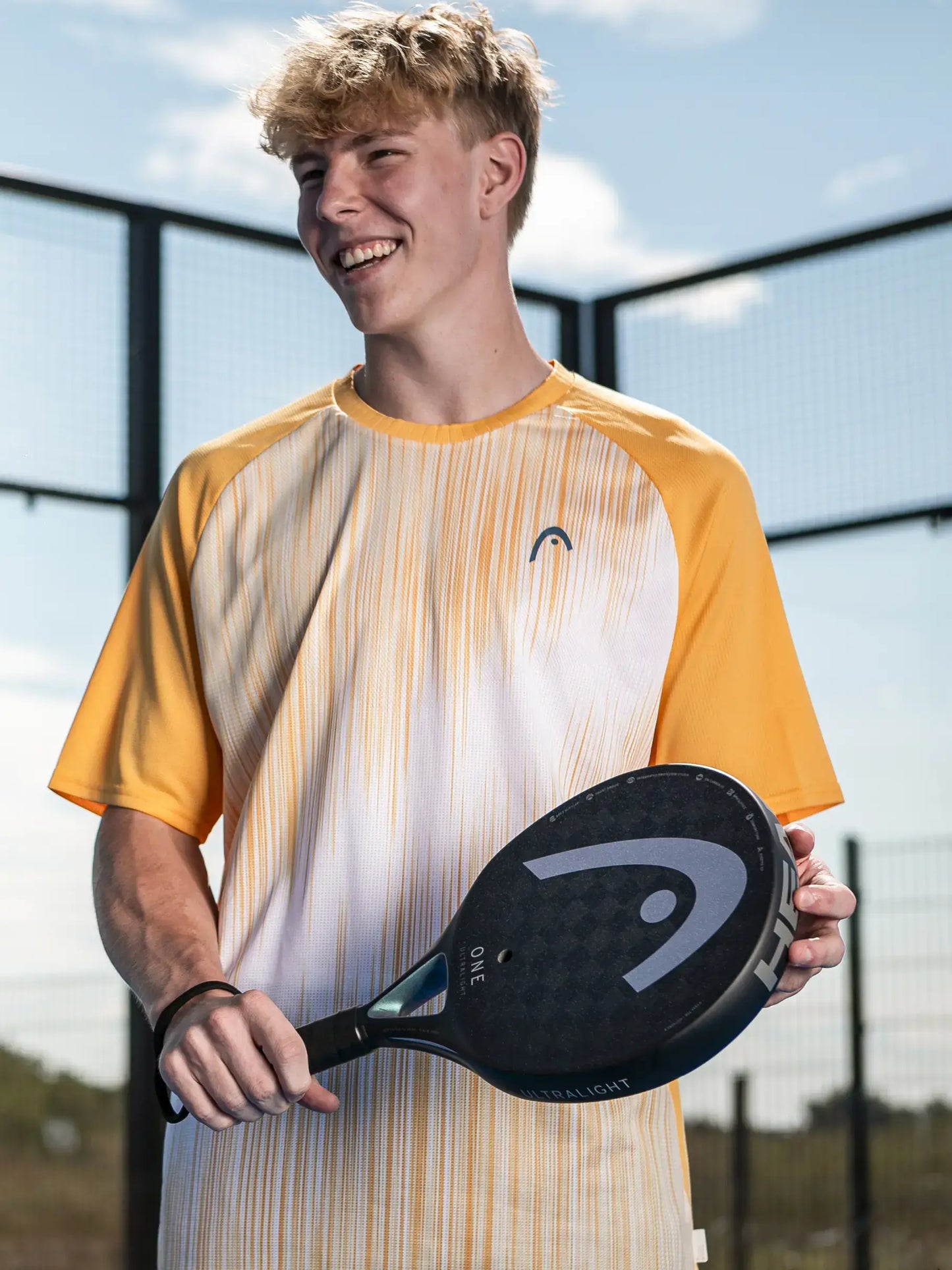 HEAD One Ultralight Padel Racquet held by a smiling player in a bright orange and white shirt during a sunny day.