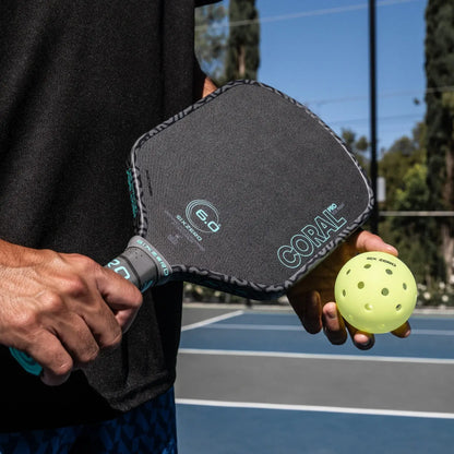 Six Zero Coral Pro 16mm paddle held with a yellow pickleball on a court background.